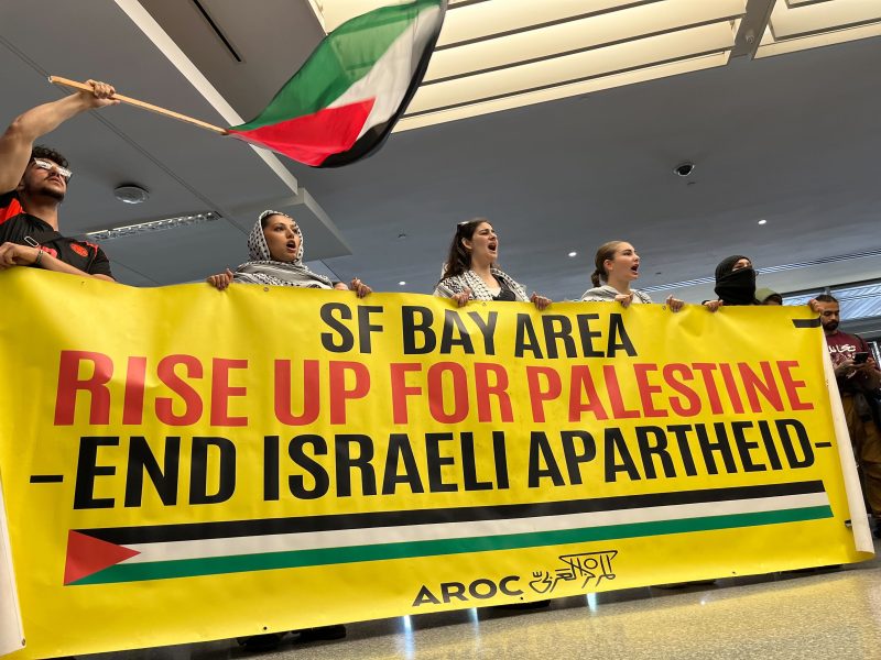 A group of people hold a banner reading "SF Bay Area Rise Up For Palestine – End Israeli Apartheid" at an indoor protest, as one person waves a Palestinian flag.