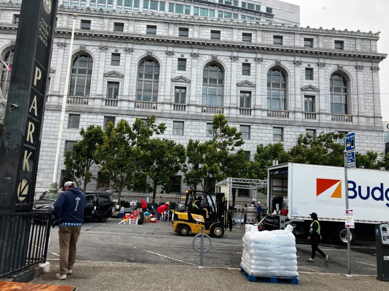 People unload supplies from trucks with pride, using a forklift in a parking lot beside a large stone building with arched windows.