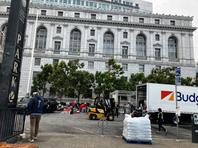 People unload supplies from trucks with pride, using a forklift in a parking lot beside a large stone building with arched windows.
