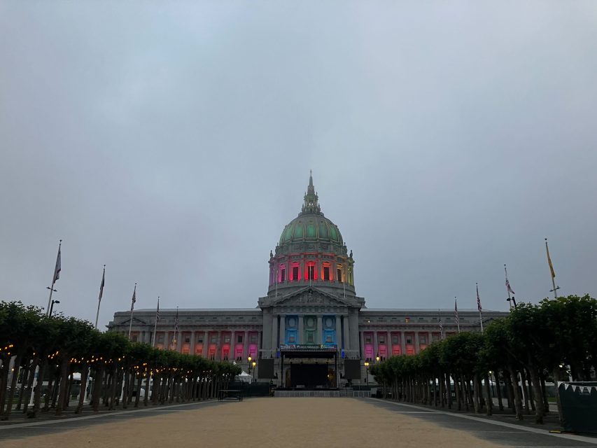 San Francisco City Hall at dusk, illuminated with pride as rainbow-colored lights glow on the dome and facade, trees and flagpoles lining the foreground.