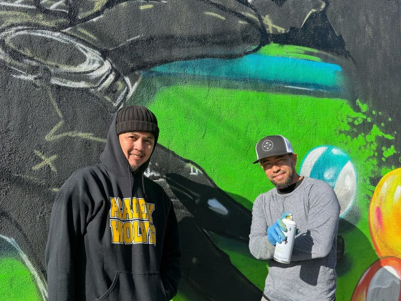 Two men pose in front of a colorful mural on a wall; one holds a spray paint can, both wear casual streetwear and hats.
