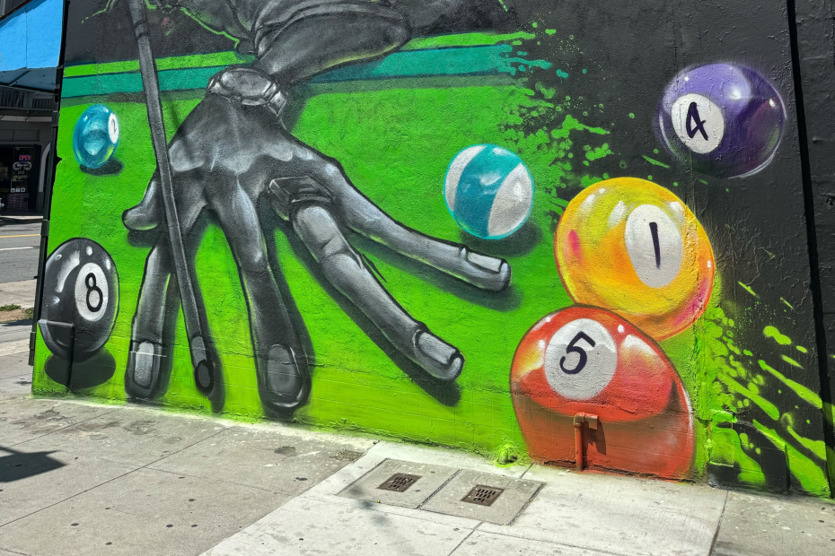 Mural of a hand holding a pool cue over a green table, with several colorful billiard balls scattered, painted on an exterior wall.