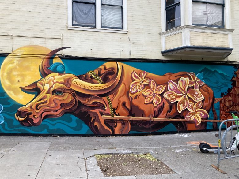 A mural of a stylized brown ox with floral patterns and a yellow moon in the background is painted on the side of a building near a sidewalk.