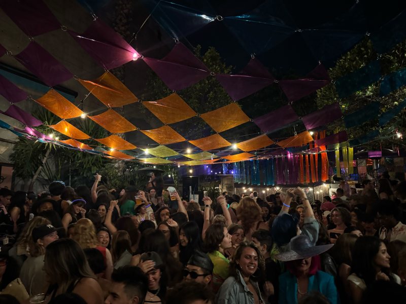A large crowd of people gather and dance at a lively outdoor pride night party under colorful, translucent fabric decorations and string lights.