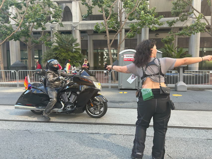 A person on a motorcycle with pride flags waits as an event staff member wearing a harness directs them along a city street, lined with barriers and onlookers celebrating pride.