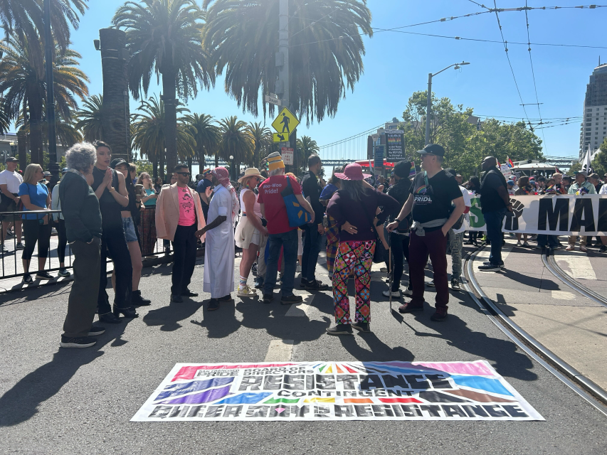 A group of people stand together behind a “Trans Resistance Contingent” banner at a pride street event, with palm trees and crowds in the background.