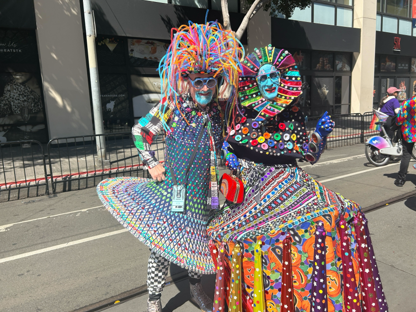 Two people in colorful, elaborate costumes with face paint pose together on a city street during a Pride parade or festival.