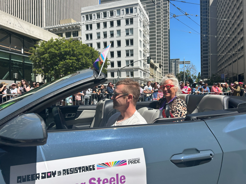 A man and a woman ride in a convertible during a pride parade, with a rainbow flag and a sign reading "QUEER JOY IS RESISTANCE" on the car door. Crowds watch from the sidewalk.