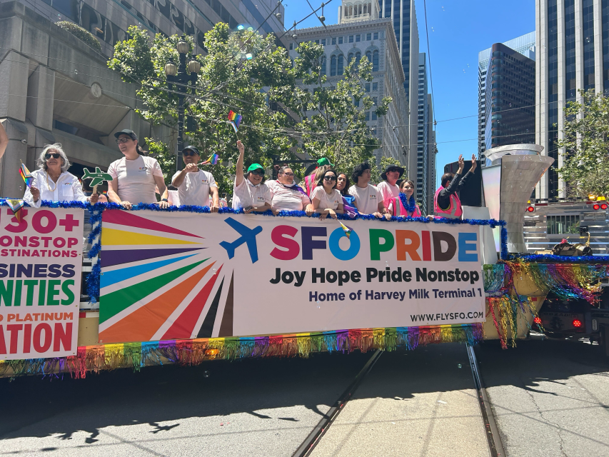 A parade float adorned with pride colors and an "SFO Pride" banner carries people in festive attire through the lively urban scene.