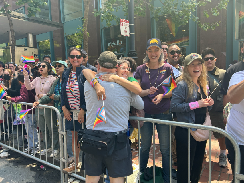 People stand behind a metal barricade at a pride parade, some waving rainbow flags. Two people in the foreground hug while others smile and watch, celebrating pride. Trees and storefronts are visible in the background.