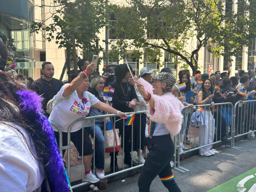 Person in a pink fur jacket interacts with a cheering crowd holding rainbow flags behind metal barricades at an outdoor pride celebration.