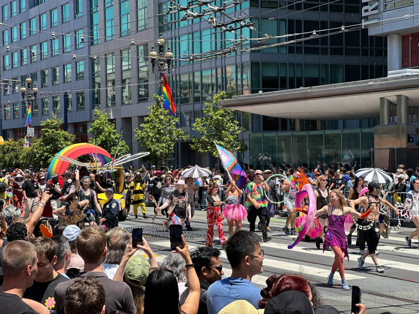 A diverse group of people in colorful costumes celebrate pride as they participate in a parade, watched by a crowd along a city street with modern buildings in the background.