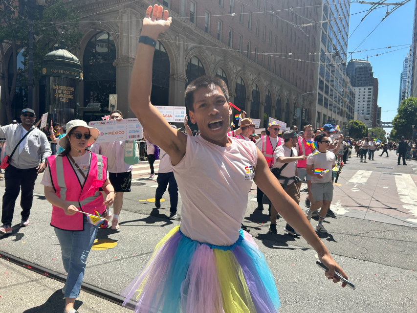 Person in a colorful tutu and white t-shirt raises an arm and smiles with pride while leading a group of people during a parade on a sunny city street.