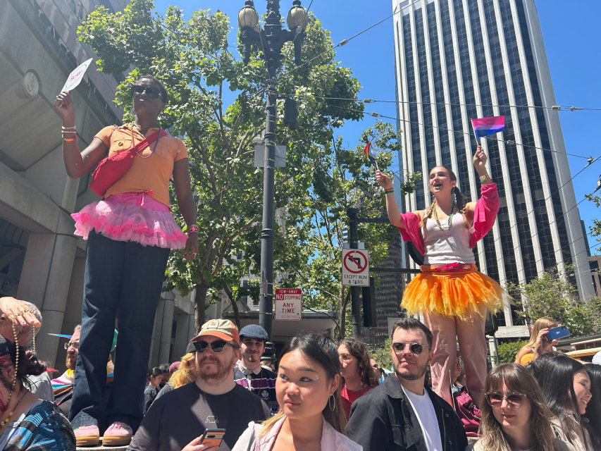 Two people in colorful tutus stand on elevated surfaces holding pride flags at an outdoor pride event, surrounded by a crowd in an urban setting with tall buildings and trees.