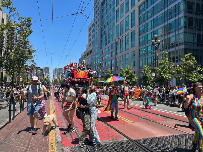 People celebrate pride in a colorful parade on a city street, some carrying rainbow flags and wearing vibrant outfits, with a decorated float and spectators lining the sidewalk.
