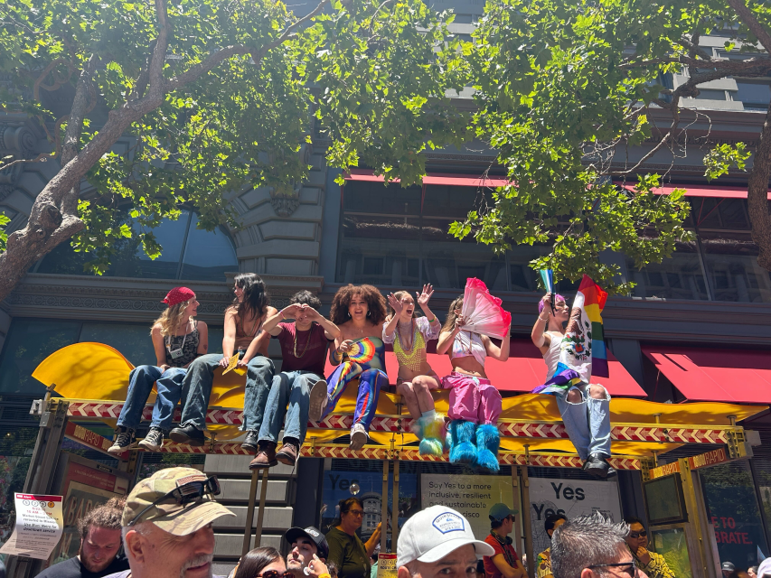 A group of people in colorful outfits sit on top of a bus stop shelter during a pride parade, surrounded by trees and sunlight, celebrating pride as the crowd gathers below.
