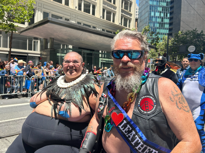 Two people participate in a pride parade, one wearing a feather collar and the other sporting a leather vest and sash that reads "LEATHER," with a crowd and city buildings in the background.