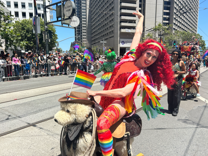 A person in a red costume with a rainbow pride flag rides a bear statue in a parade, surrounded by spectators and city buildings.