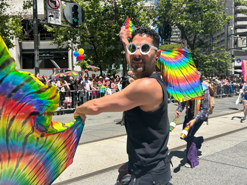 A person wearing white sunglasses and a black tank top holds rainbow-colored pride wings while performing in a street parade before a crowd.