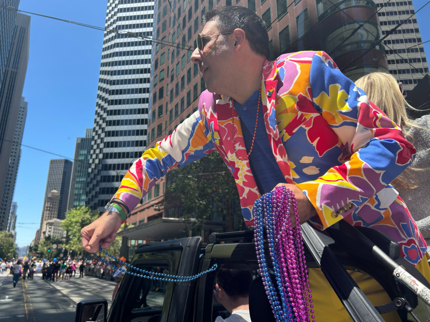 A person in a colorful floral jacket leans out of a vehicle at a pride parade, handing out vibrant bead necklaces on a city street surrounded by tall buildings.