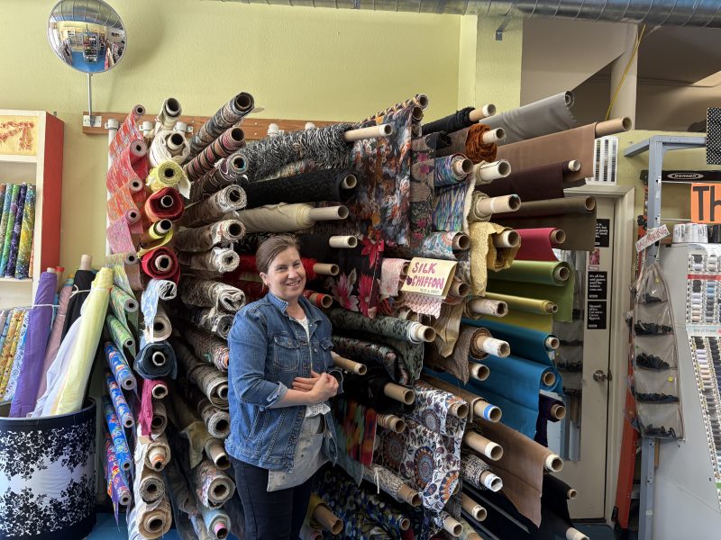 A woman stands and smiles in front of shelves filled with various colorful rolls of fabric inside a fabric store.