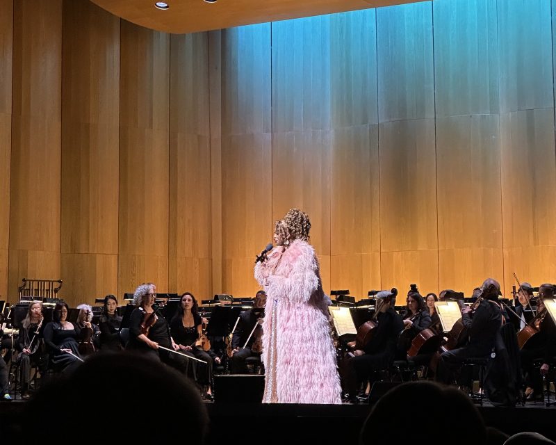 A performer in a pink feathered outfit sings onstage in front of an orchestra, with musicians seated and playing string instruments.