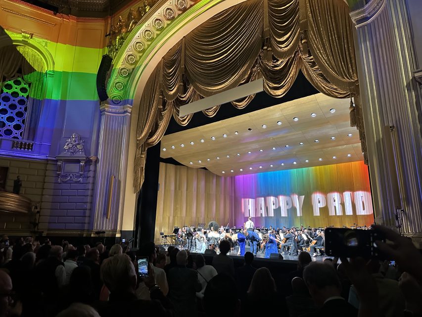 An orchestra performs on stage under gold curtains with "HAPPY PRIDE" and rainbow lights projected on the back wall; audience members watch and take photos.