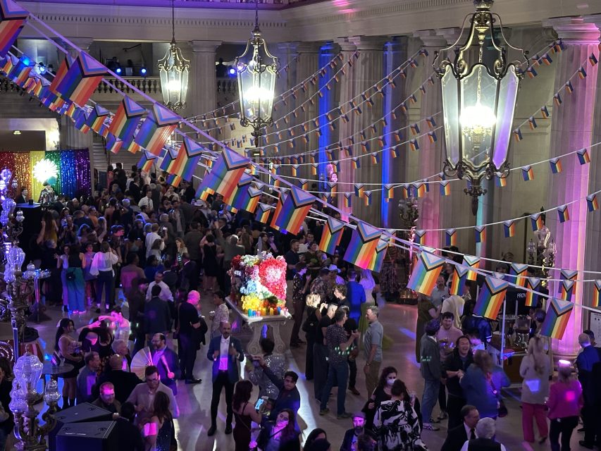 Large indoor event with many people socializing under hanging Progress Pride flags in a grand hall with columns and chandeliers. Colorful lights illuminate the festive scene.