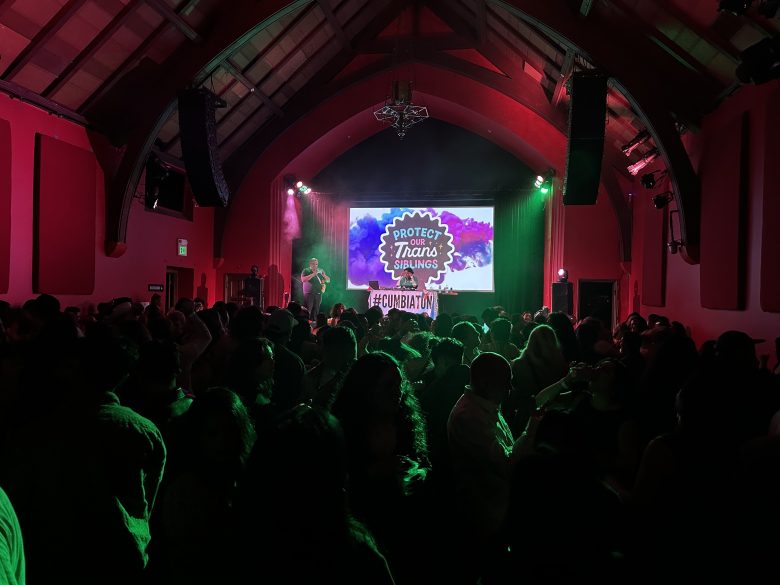 A crowd gathers at an indoor Pride event with a stage, where a screen displays the message “Protect Trans Siblings” and a #CUMBIATON hashtag. Green lighting fills the room.