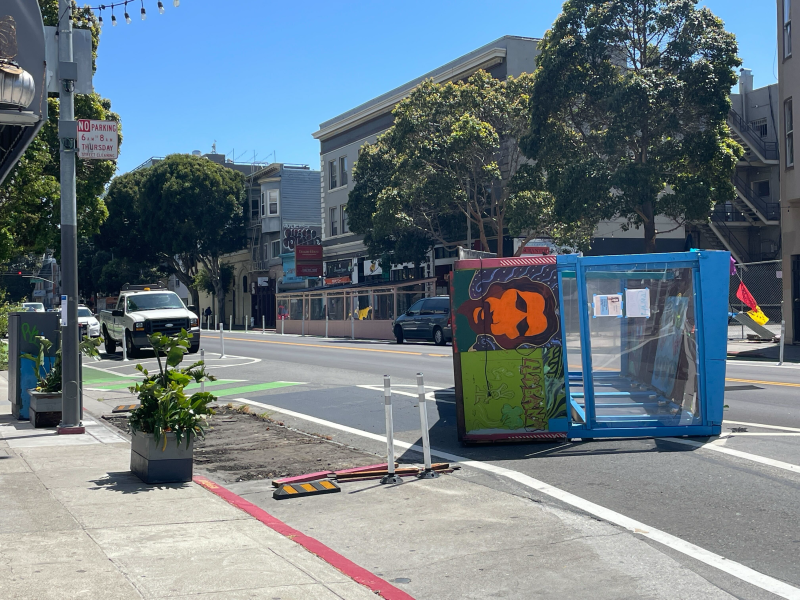 A colorful painted structure is tipped over on a city street near the sidewalk; a city vehicle is parked nearby and trees line the road.