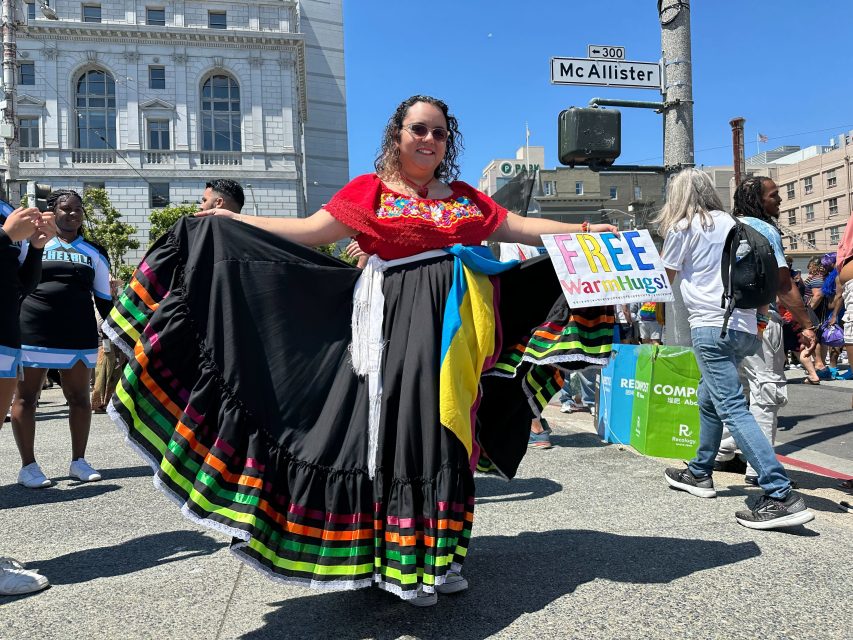 A person in a colorful traditional dress stands on a city street during a pride event, holding a sign that says "FREE warm hugs!" and spreading positivity to passersby.
