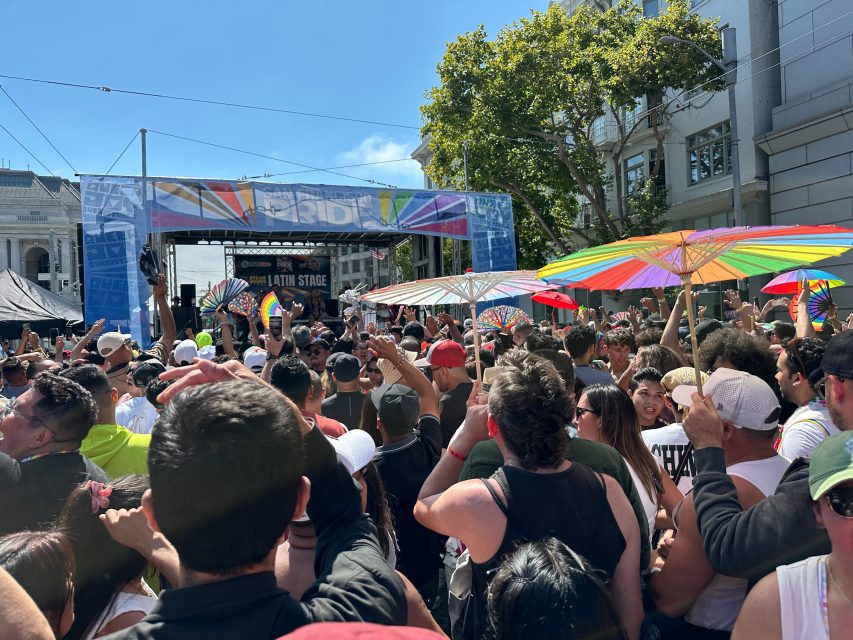 A large crowd gathers in front of an outdoor stage at a pride festival, with several people holding rainbow-colored umbrellas and fans under a sunny sky.