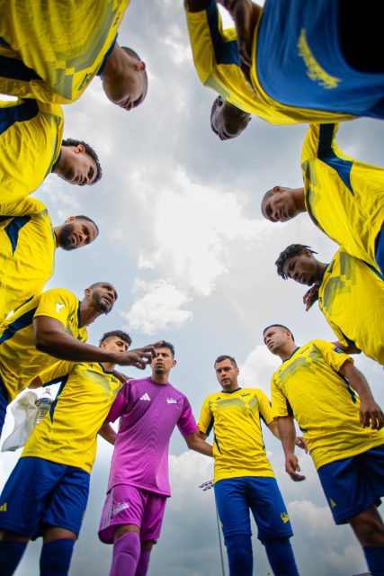 Soccer players in yellow jerseys and a goalkeeper in pink huddle together on the field, preparing for a game under a cloudy sky.