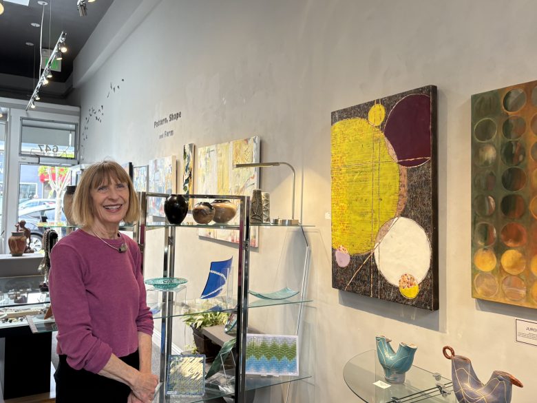 A woman stands beside glass art and paintings displayed on shelves and walls in a well-lit gallery.