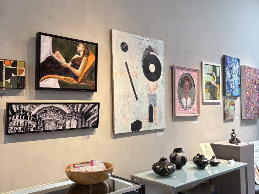 A gallery wall displays various artworks, including paintings and photographs, above a shelf with black pottery, a sculpture, and a bowl with small items.