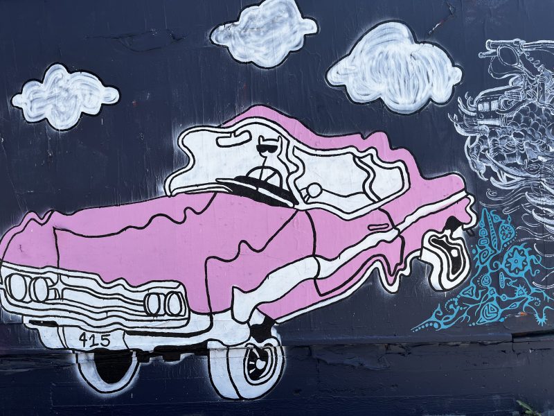 A mural of a pink classic car outlined in white, with clouds above it and decorative blue patterns near the rear tire, painted on a dark wall.