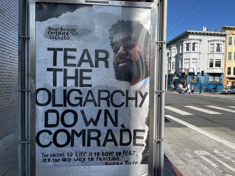 A street poster on a bus stop reads, "TEAR THE OLIGARCHY DOWN, COMRADE." with a man's face behind the text and a city street with buildings and rainbow flags in the background.