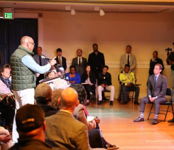 A man speaks at a podium to an audience in a conference room; several people, including media, are seated or standing, and a presentation screen is visible.
