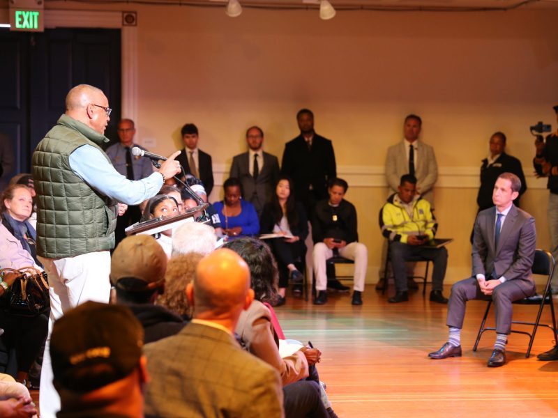 A man speaks at a podium to an audience in a conference room; several people, including media, are seated or standing, and a presentation screen is visible.