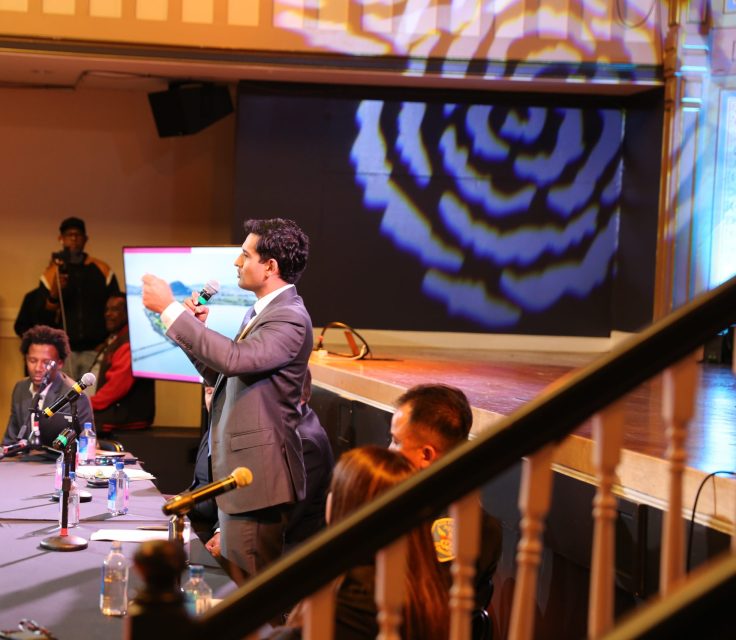 A man in a suit speaks at a panel discussion in a large room with an audience, while others sit at a table with microphones and papers.