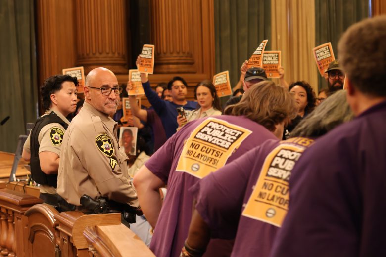 A group of people wearing purple shirts hold signs that read "Protect Public Services: No Cuts! No Layoffs!" as uniformed officers stand nearby in a large wood-paneled room.