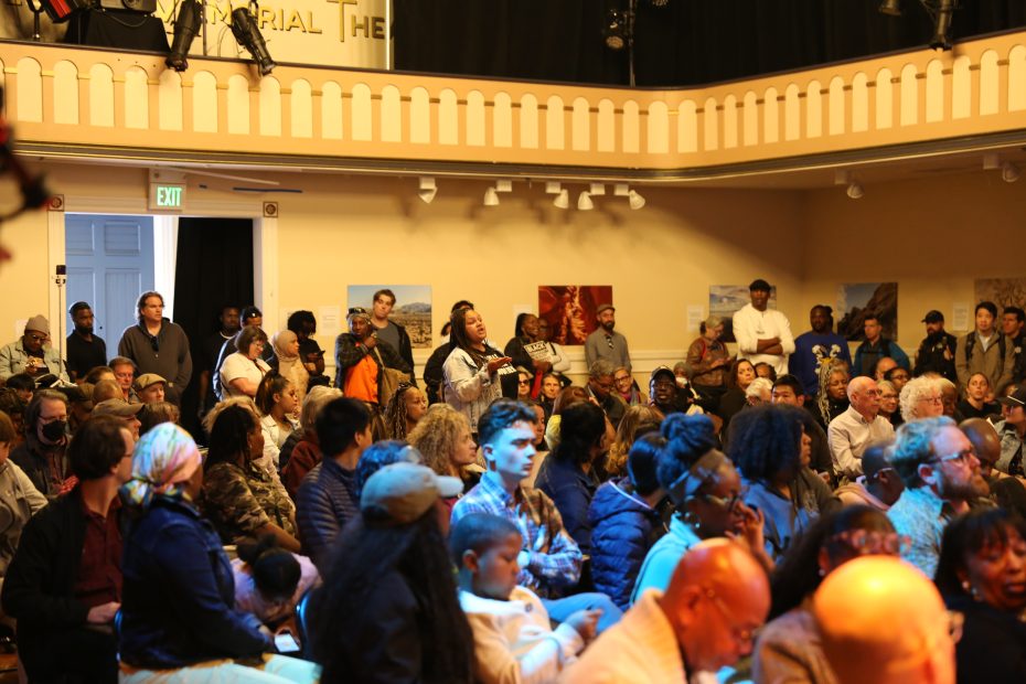 A diverse group of people sit and stand in a crowded auditorium, attentively listening to a speaker off camera.