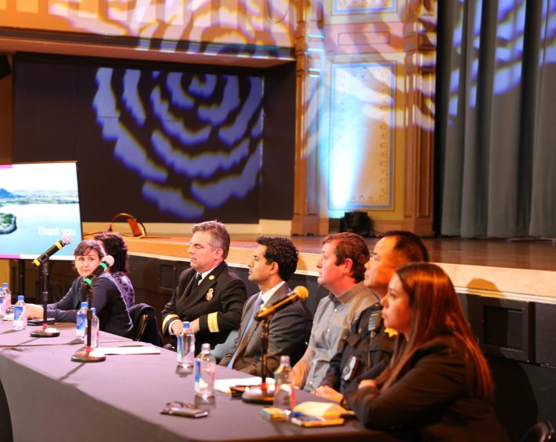 A group of seven people sit at a long table with microphones on a stage, participating in a panel discussion in a well-lit room.