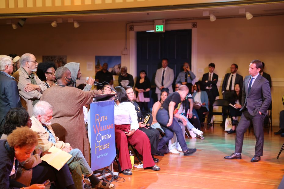 A community member speaks at a microphone in a crowded auditorium during a public meeting at Ruth Williams Opera House while others listen and a man in a suit stands in front.
