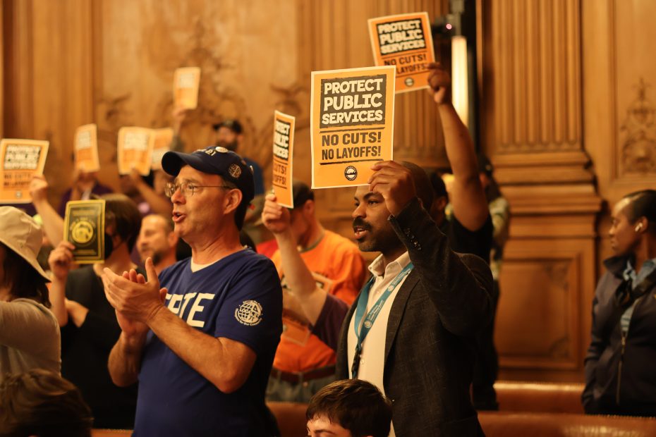 A group of people indoors hold up signs that read "Protect Public Services, No Cuts! No Layoffs!" during a gathering or protest.