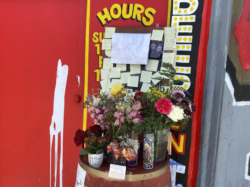 A small memorial with flowers, candles, photos, and handwritten notes displayed on a barrel against a red wall with business hours painted beside it.