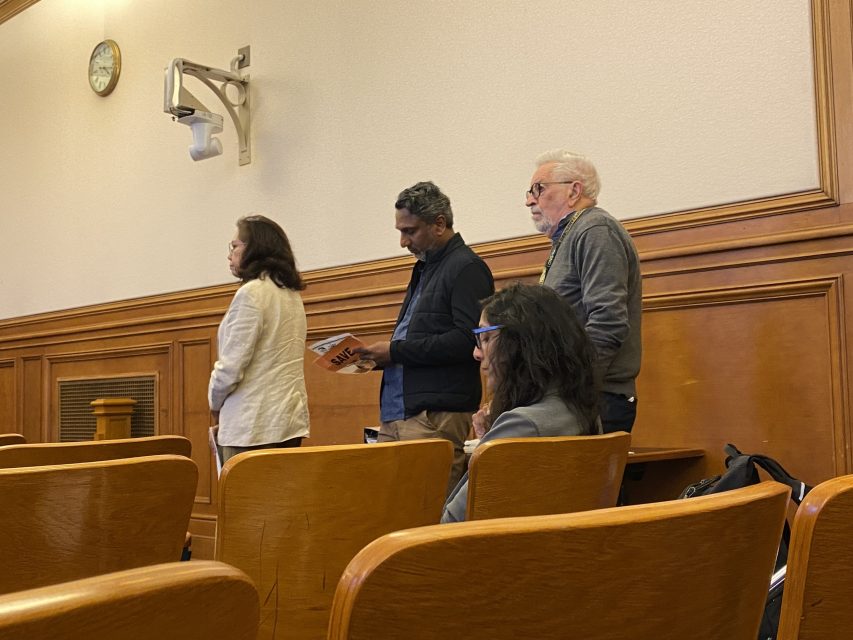 Three people stand near the wall of a courtroom, while two people sit in the wooden benches. A clock and light fixture are mounted on the wall.