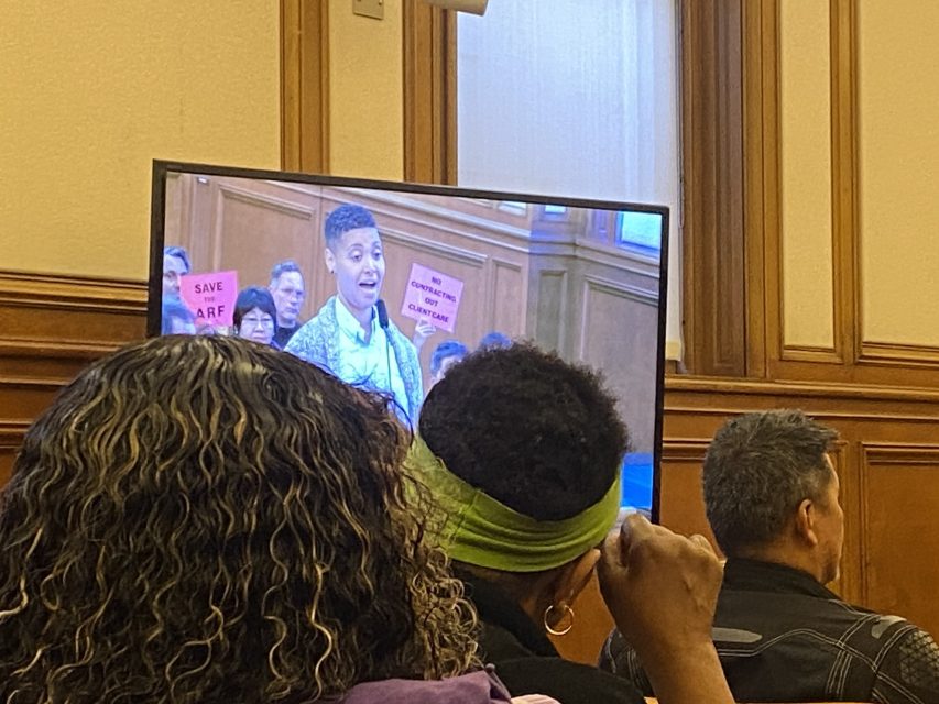 Jennifer  speaks at a public meeting shown on a screen, while audience members watch and hold pink signs that read "SAVE ARF.