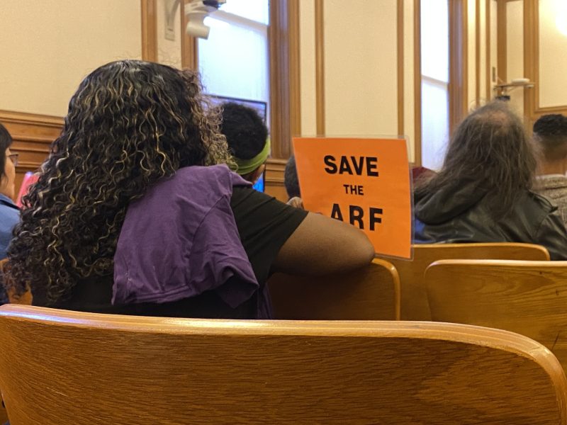A person sits in a wooden bench holding an orange sign that reads "SAVE THE ARF" in a room with large windows and several other seated people.