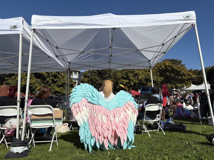 Person wearing large pastel pink and blue wings sits under a white canopy among others on folding chairs at an outdoor event.